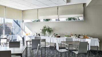 Un salón de conferencias con mesas y sillas, una planta grande y un ventanal con persianas.