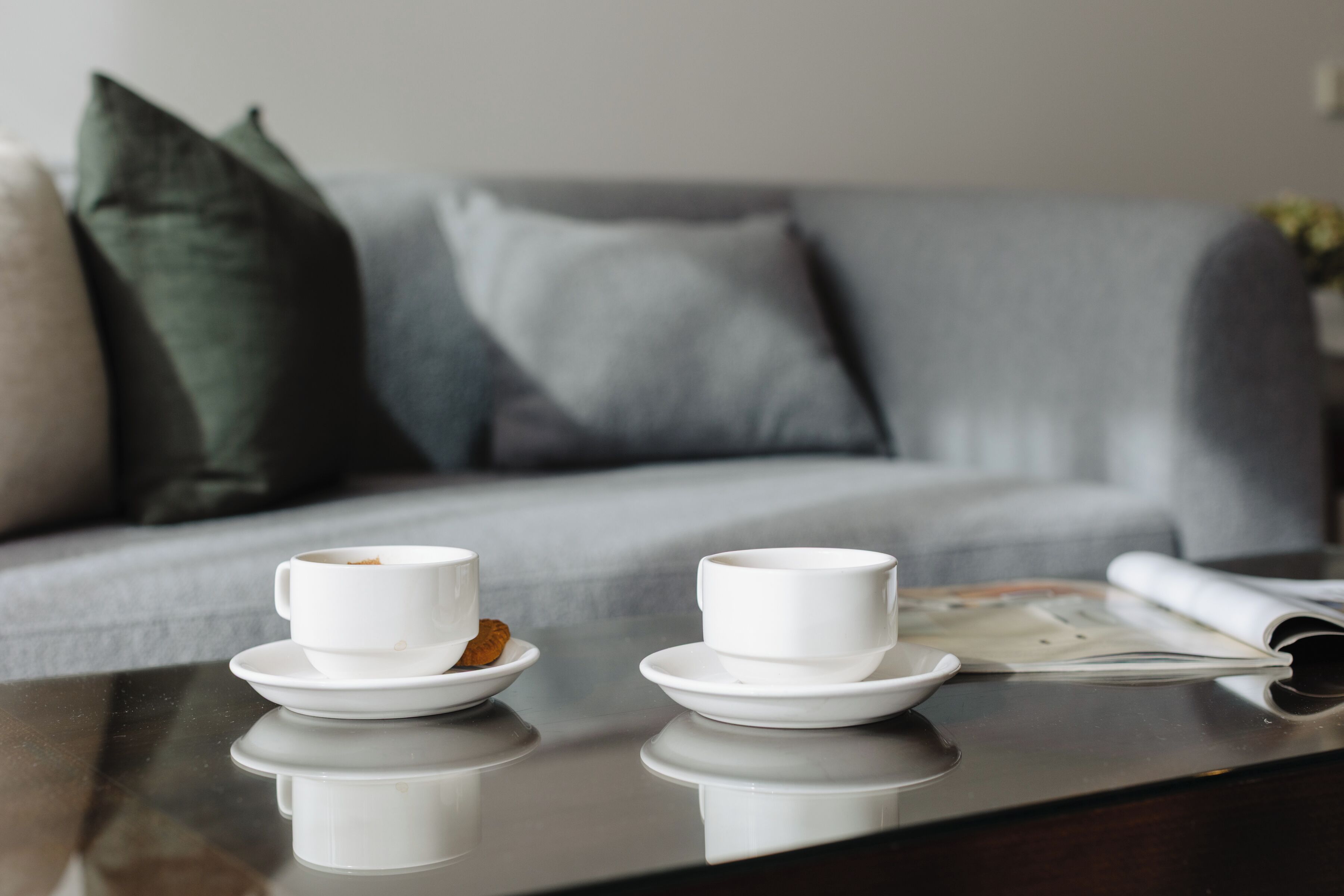 A modern living room with a glass coffee table, two white cups on saucers, and a magazine.