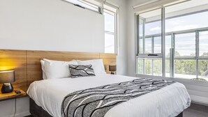 Une chambre d’hôtel moderne dotée d’un grand lit, d’une tête de lit en bois, d’une table de chevet avec une lampe et une plante, et d’une grande fenêtre donnant sur l’extérieur.