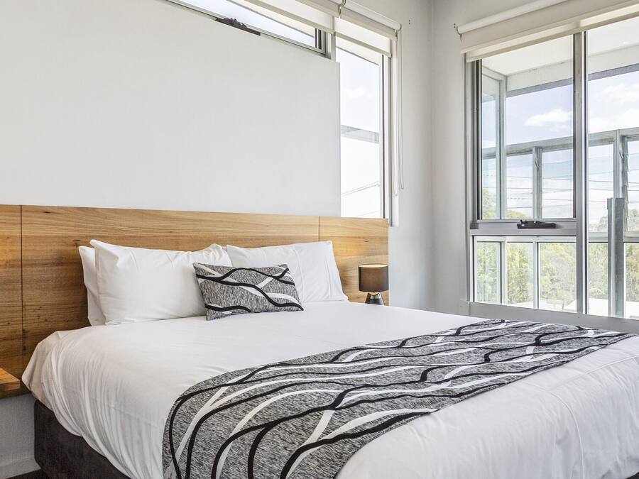 Una habitación de hotel moderna con una cama grande, un cabecero de madera, una mesita de noche con una lámpara y una planta, y un ventanal con vistas al exterior.