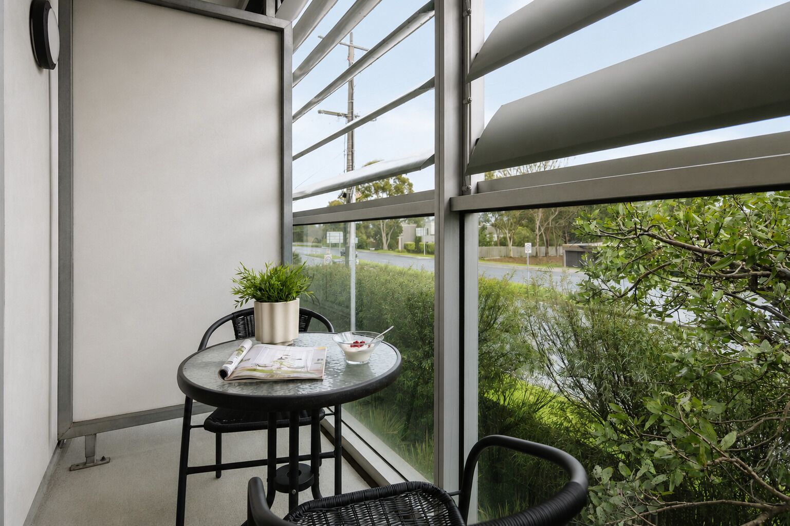 Un balcon avec une table ronde, deux chaises, un pot de fleurs, un bol avec une cuillère et un journal.