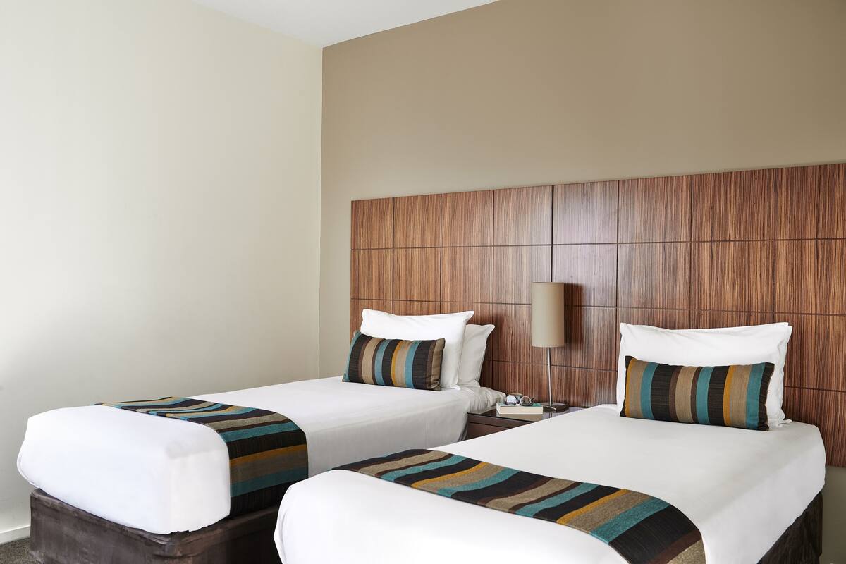 Habitación de hotel con dos camas, cabecero de madera, mesita de noche con lámpara y manta a rayas.