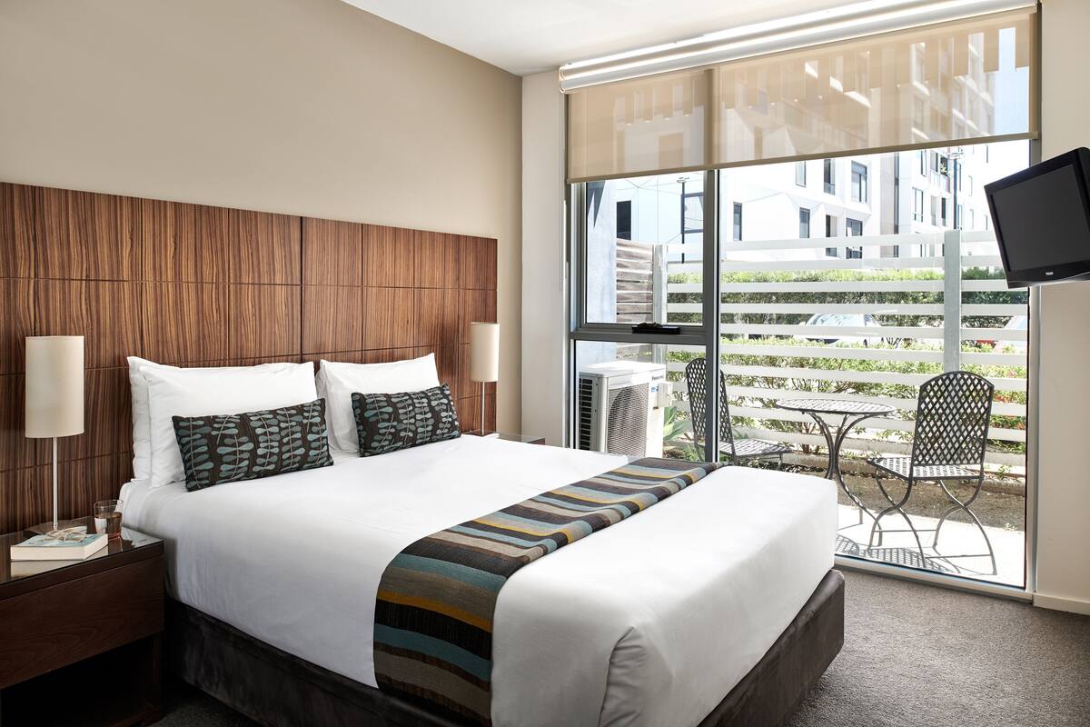 Una habitación de hotel con una cama grande, dos mesitas de noche, un televisor y un balcón con mobiliario exterior.