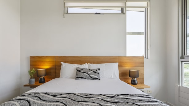 Un lit bien fait, recouvert d’une couvre-lit à motifs, avec deux lampes de chevet et une petite plante en pot posée sur une table de chevet.