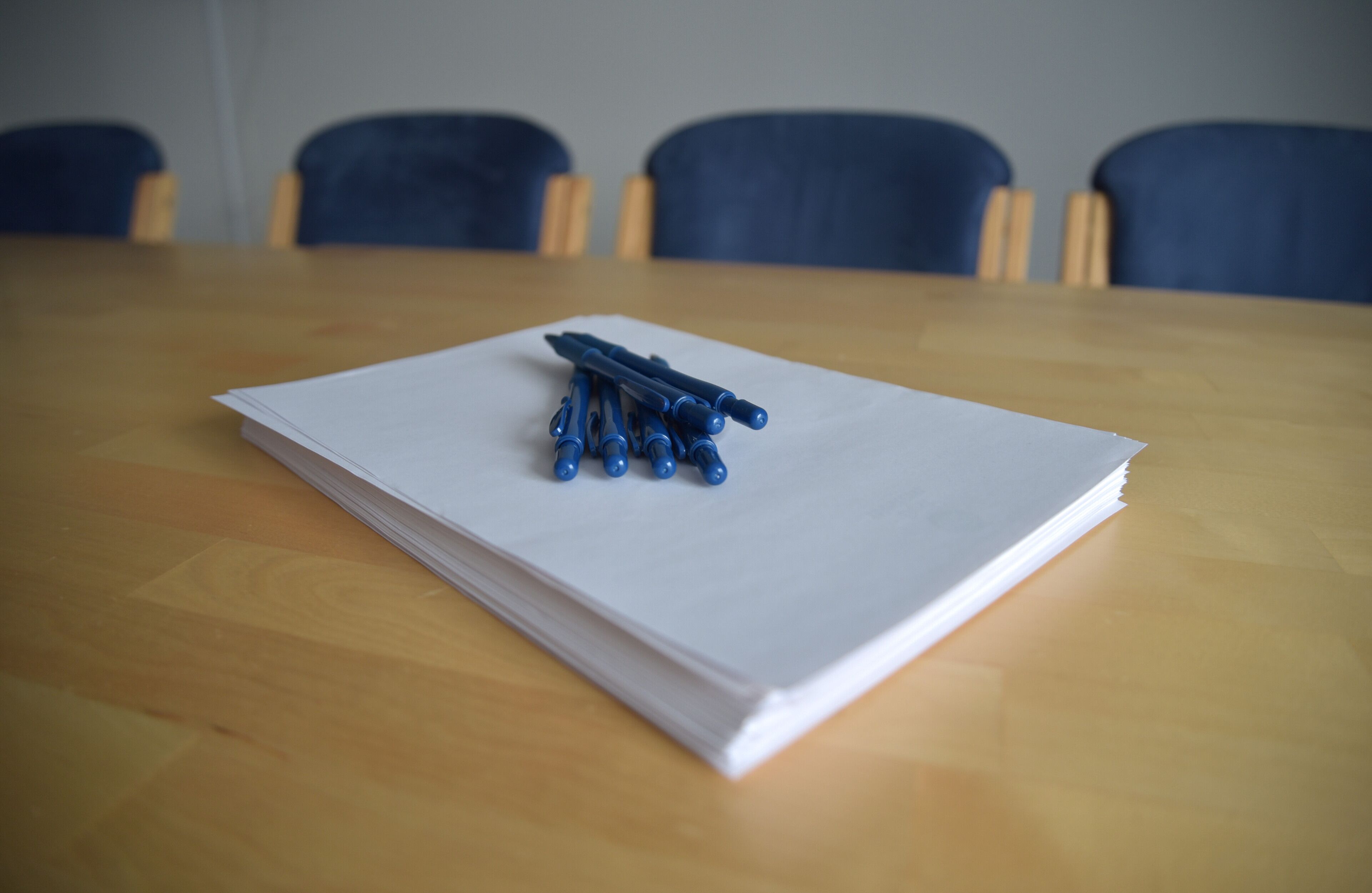 A stack of white papers on a wooden table with blue pens arranged on top.