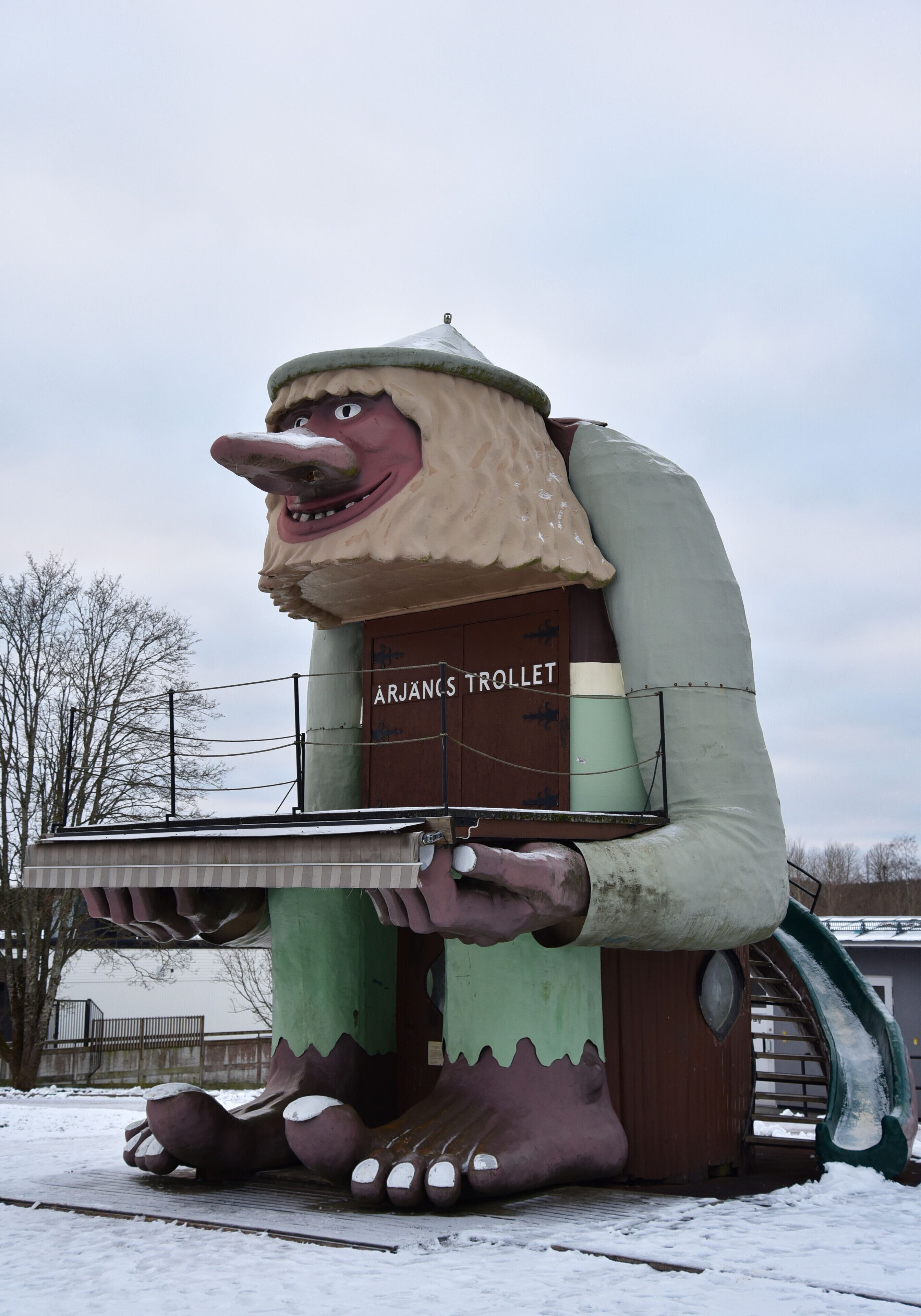 A large, whimsical statue of a character with a bucket, labeled "ÁRJÁNS TROLLET," in a snowy outdoor setting.