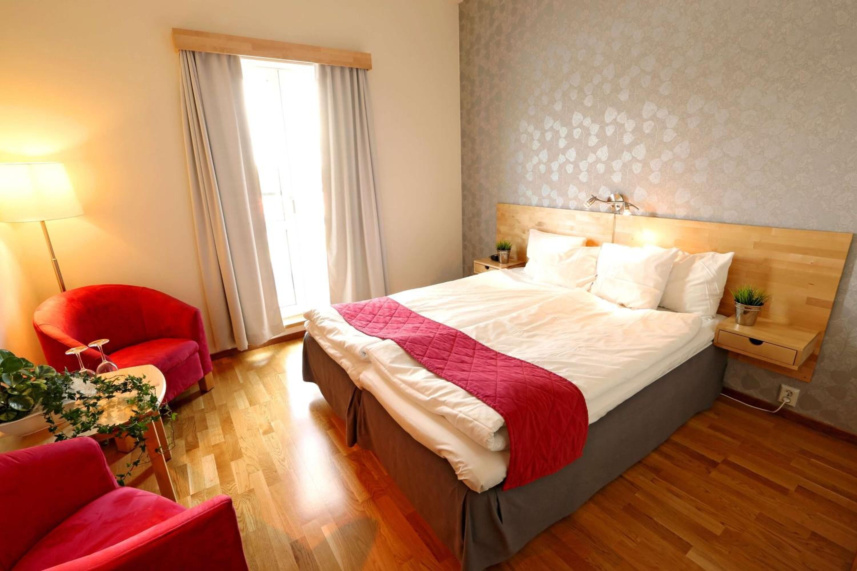 A bedroom with a wooden bed, a red chair, a bedside table with a lamp, and a window with curtains.