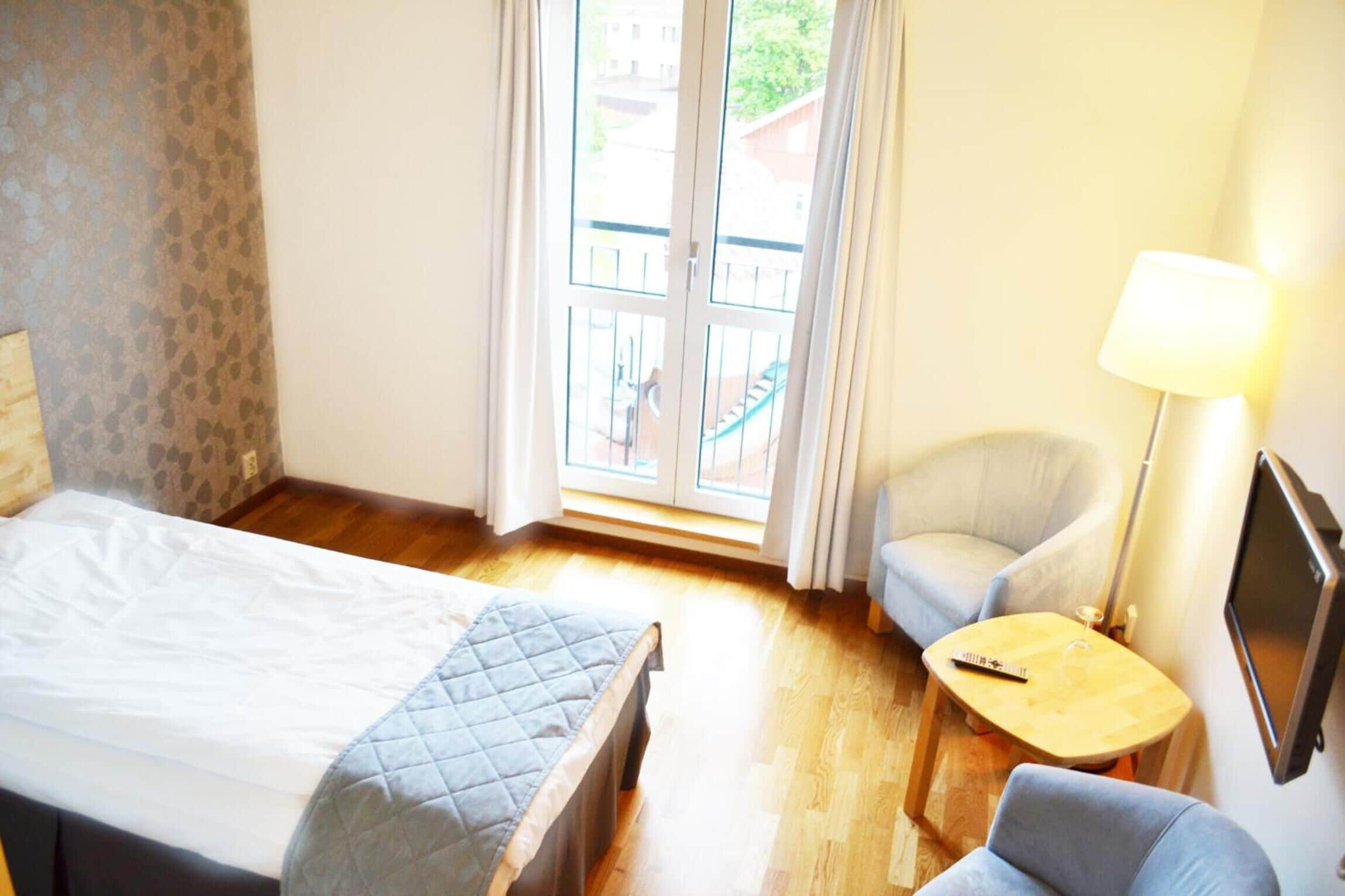 Une chambre d’hôtel comprenant un lit, une chaise, une petite table, un téléviseur et un balcon offrant une vue sur les bâtiments et la verdure.