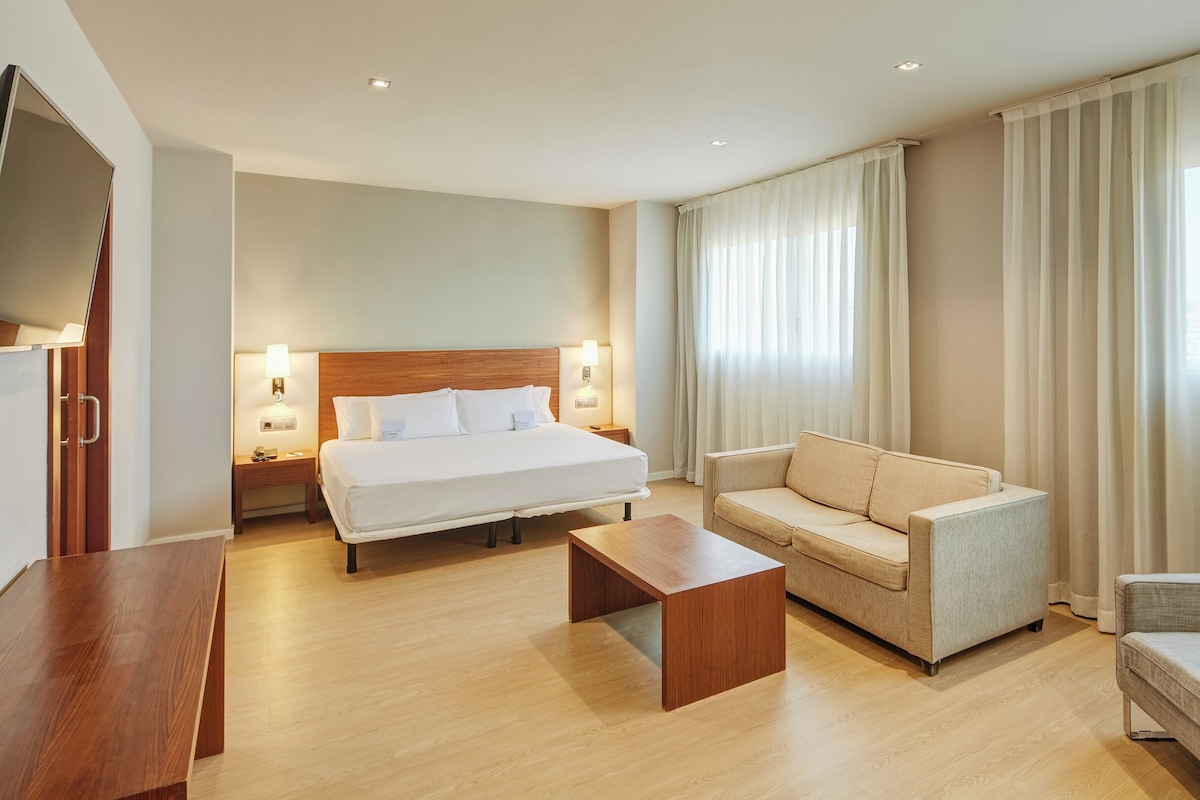 A hotel room with a bed, a sofa, a chair, a wooden table, and a TV.