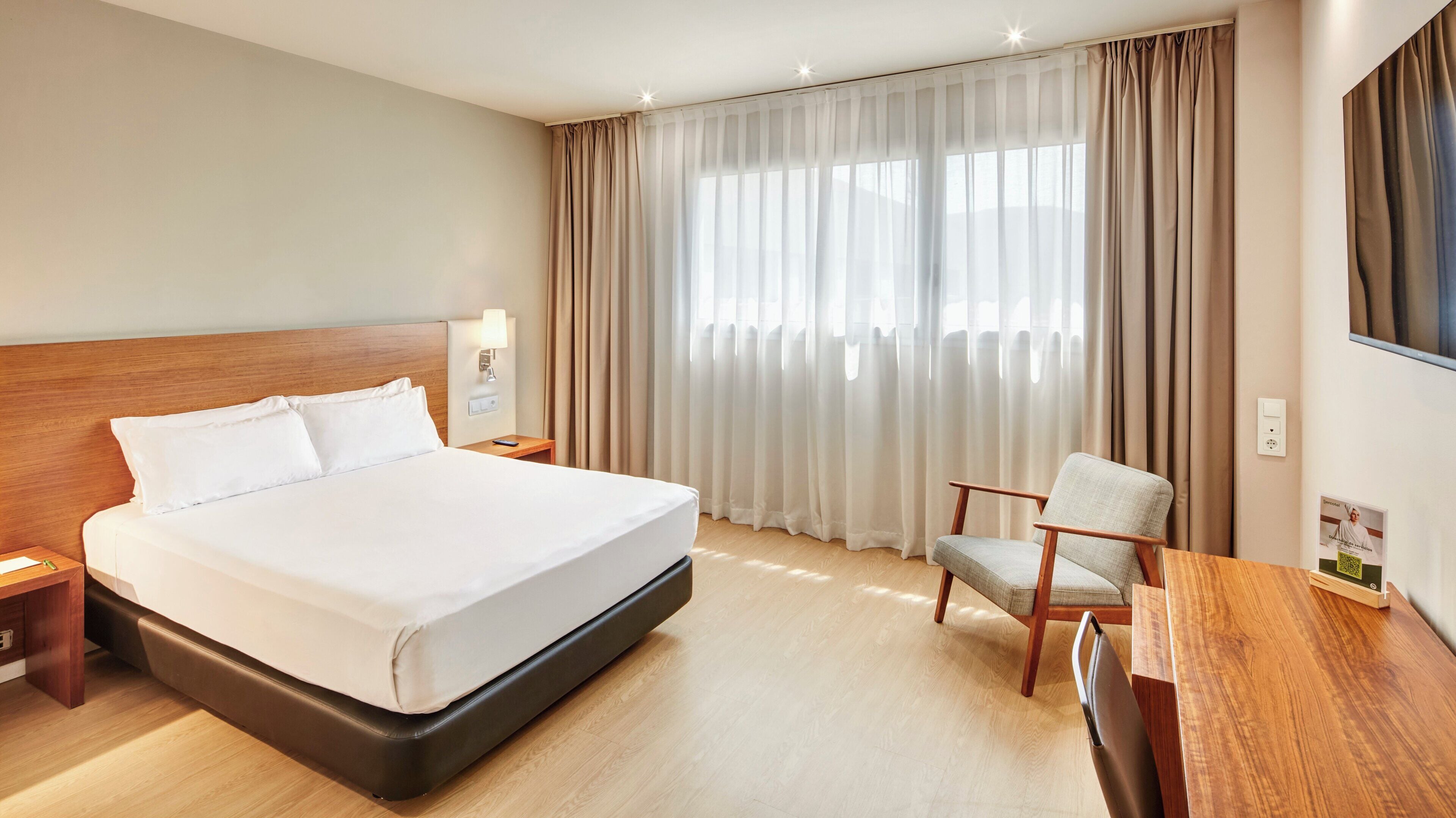 A hotel room with a large bed, a wooden nightstand, a desk with a chair, and a large window with curtains.