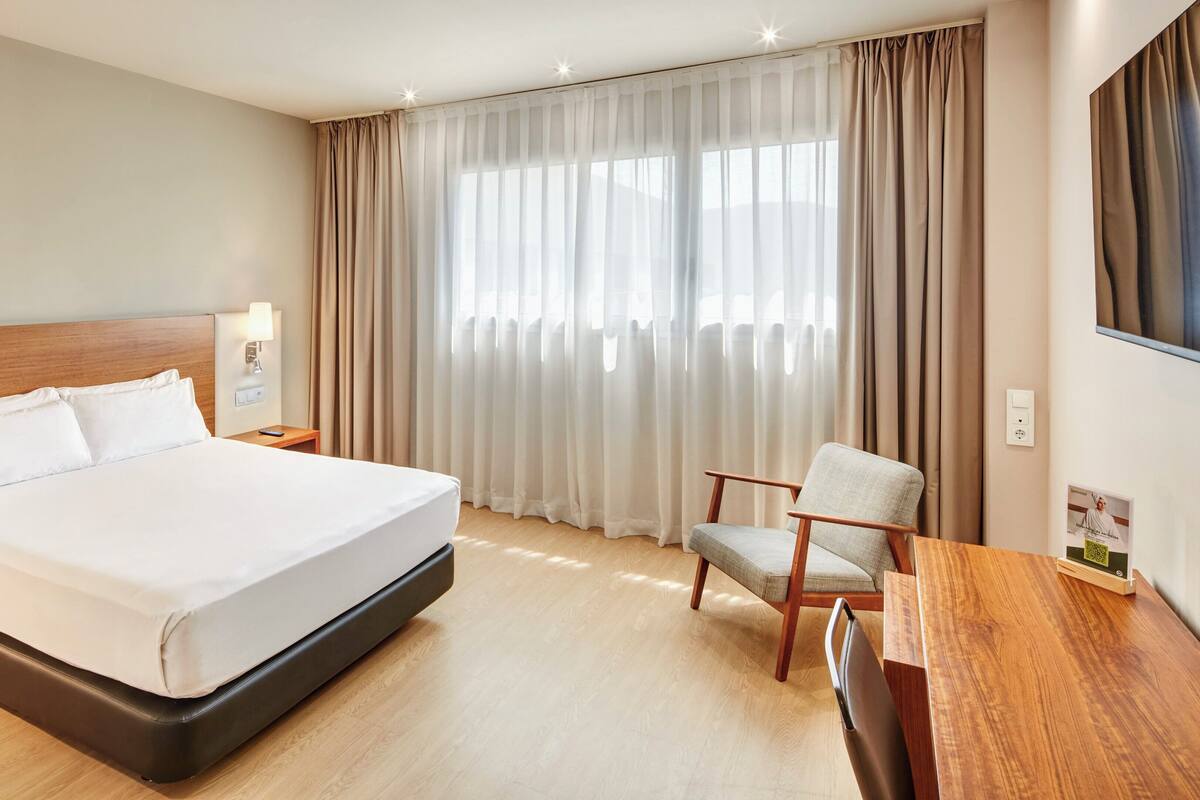 Hotellihuone, jossa on suuri sänky, puinen yöpöytä, työpöytä tuolilla sekä suuri ikkuna, jossa on verhot.