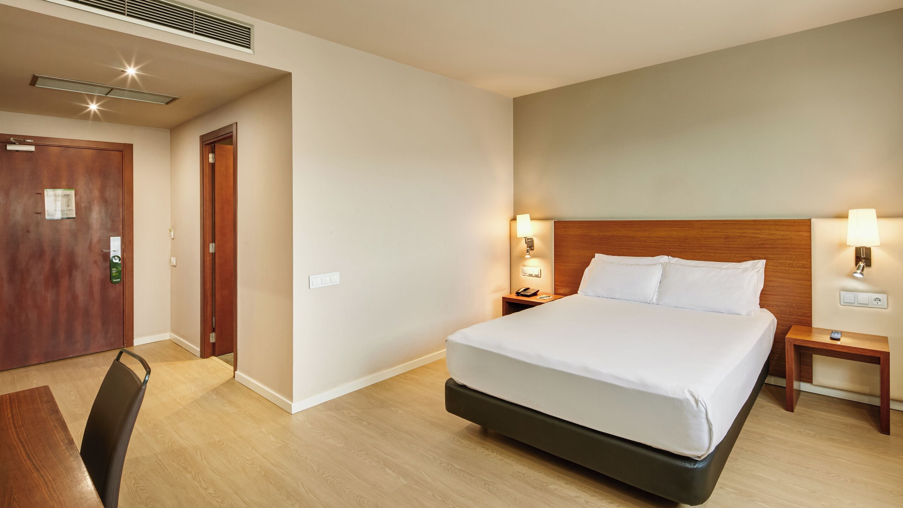 Habitación de hotel con una cama grande, cabecera de madera, lámparas de noche, una mesita de noche de madera y un escritorio con una silla.