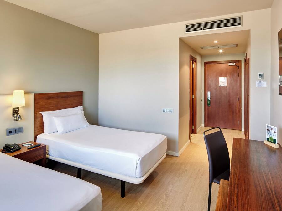 Habitación de hotel con dos camas individuales, una mesita de noche con lámpara, un escritorio con silla y un espejo grande en la pared.
