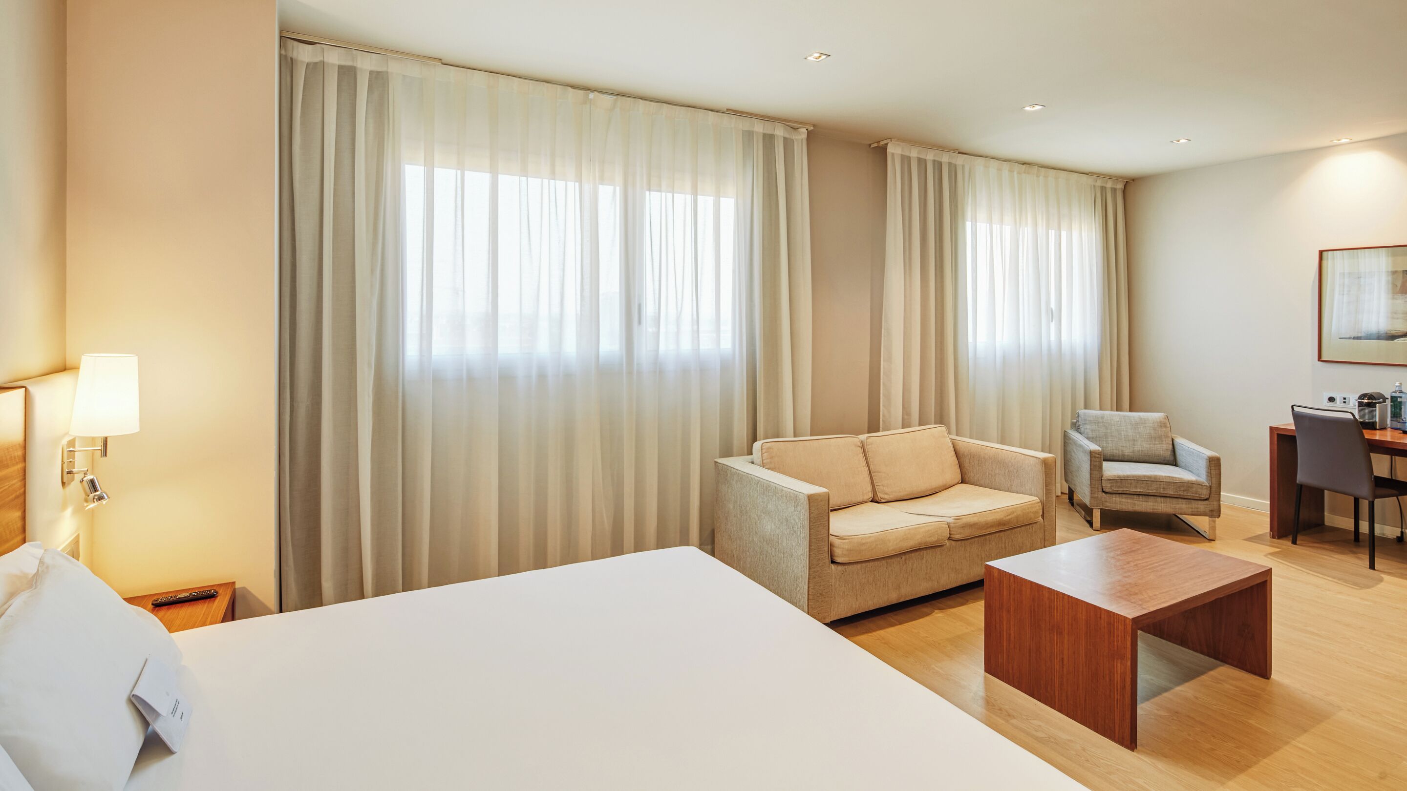 A hotel room with a large bed, two armchairs, a small table, and a desk with a lamp.