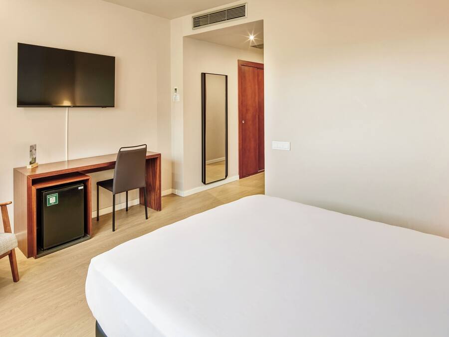 A hotel room with a bed, a desk with a chair, a television, a mirror, and a door.