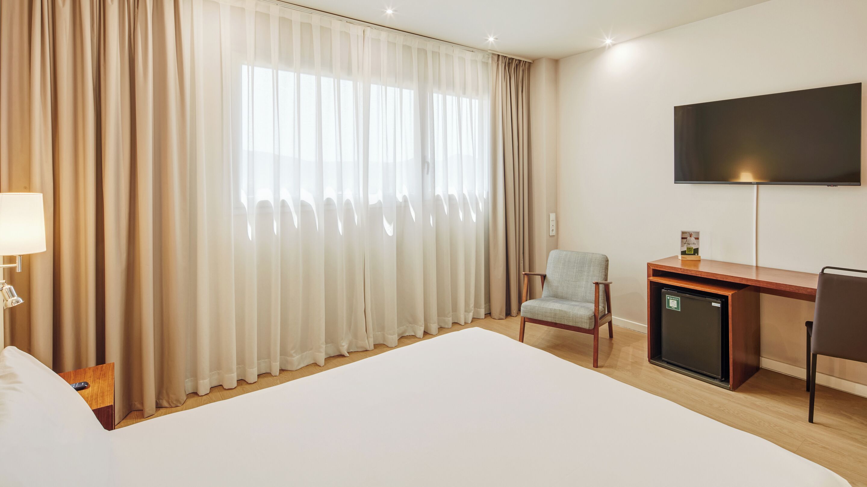 A hotel room with a large bed, a desk with a computer, a chair, a TV mounted on the wall, and a window with curtains.