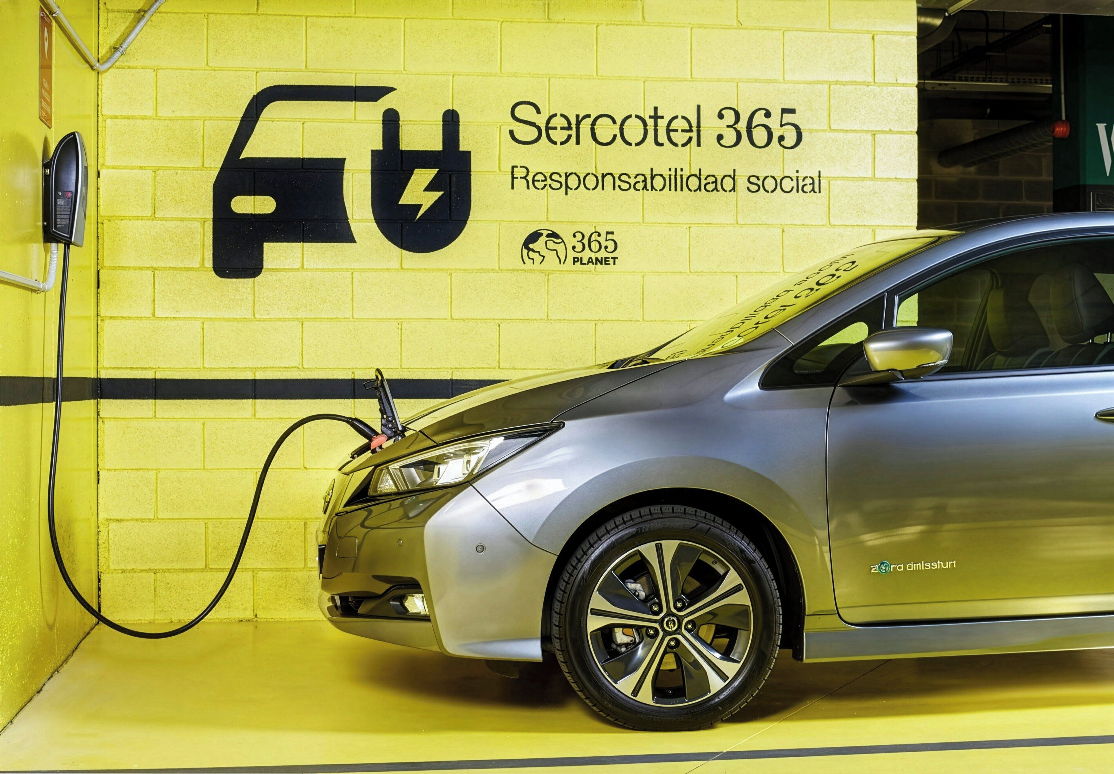 Un auto plateado cargándose en una estación con un muro de ladrillos amarillos, un letrero que dice 'Sercotel 365 Responsabilidad social' y el logo de '365 Planet'.