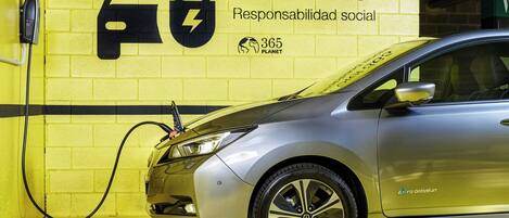 Un auto plateado cargándose en una estación con un muro de ladrillos amarillos, un letrero que dice 'Sercotel 365 Responsabilidad social' y el logo de '365 Planet'.