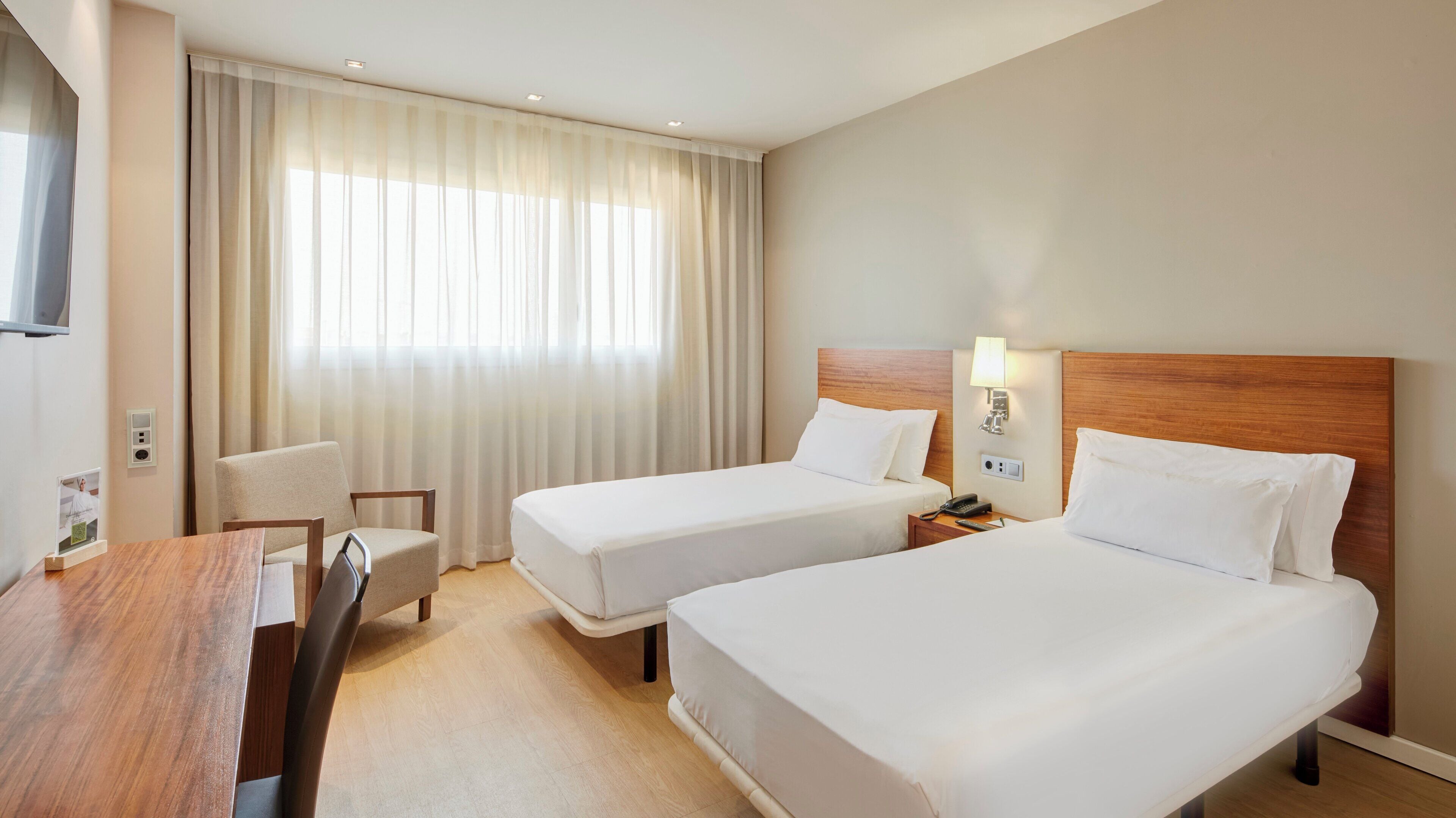 A hotel room with two beds, a desk, a chair, a TV, and a window with curtains.