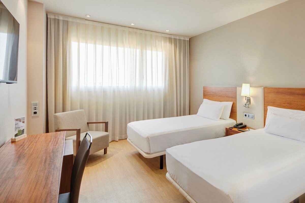 Habitación de hotel con dos camas, un escritorio, una silla, un televisor y una ventana con cortinas.