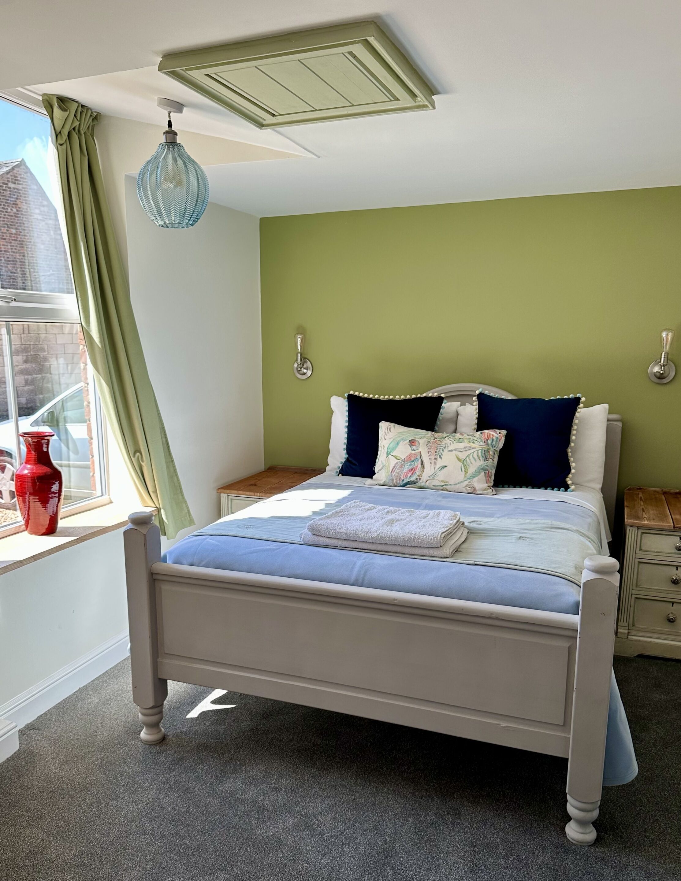 Un dormitorio con cama, una ventana con cortinas, un jarrón rojo y un respiradero en el techo.