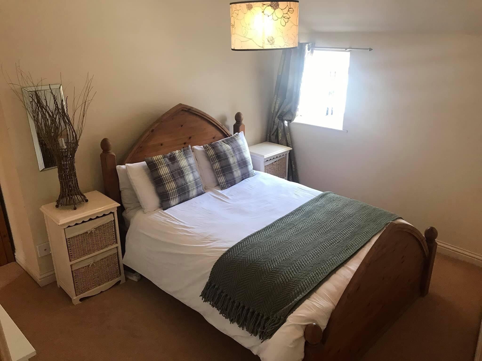 Ein ordentlich bezogenes Bett mit Holz-Kopfteil, ein Nachttisch mit Lampe und ein Fenster mit Vorhängen.