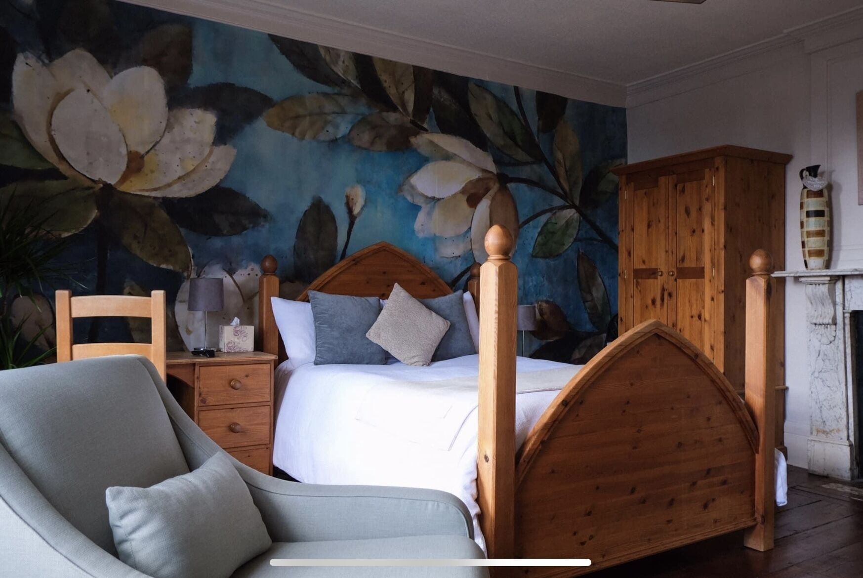 A bedroom with a large floral mural, a wooden four-poster bed, a nightstand with a lamp, and a chair.