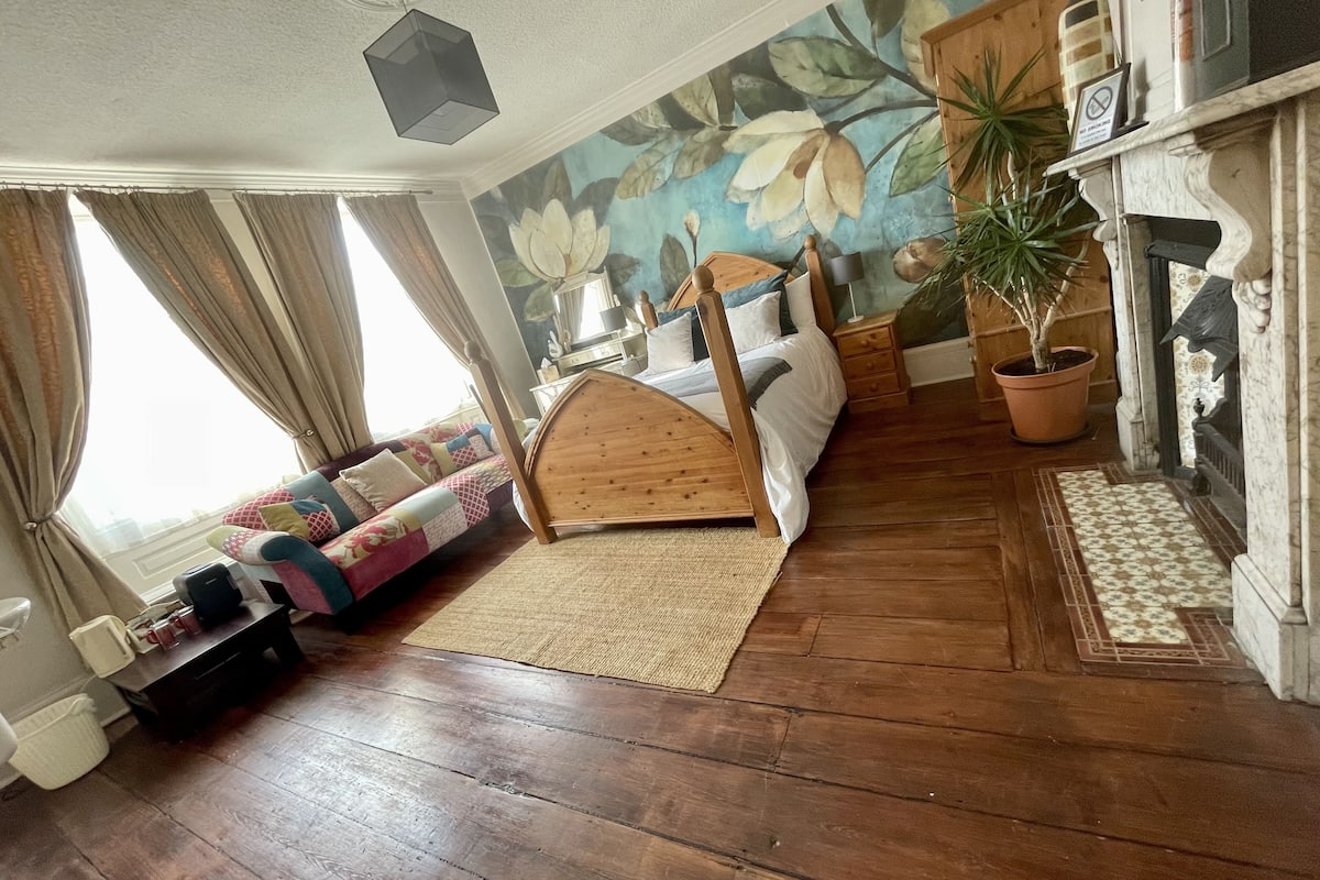 Un dormitorio acogedor con una cama de madera, un sofá, un ventanal con cortinas, una chimenea y un mural floral.