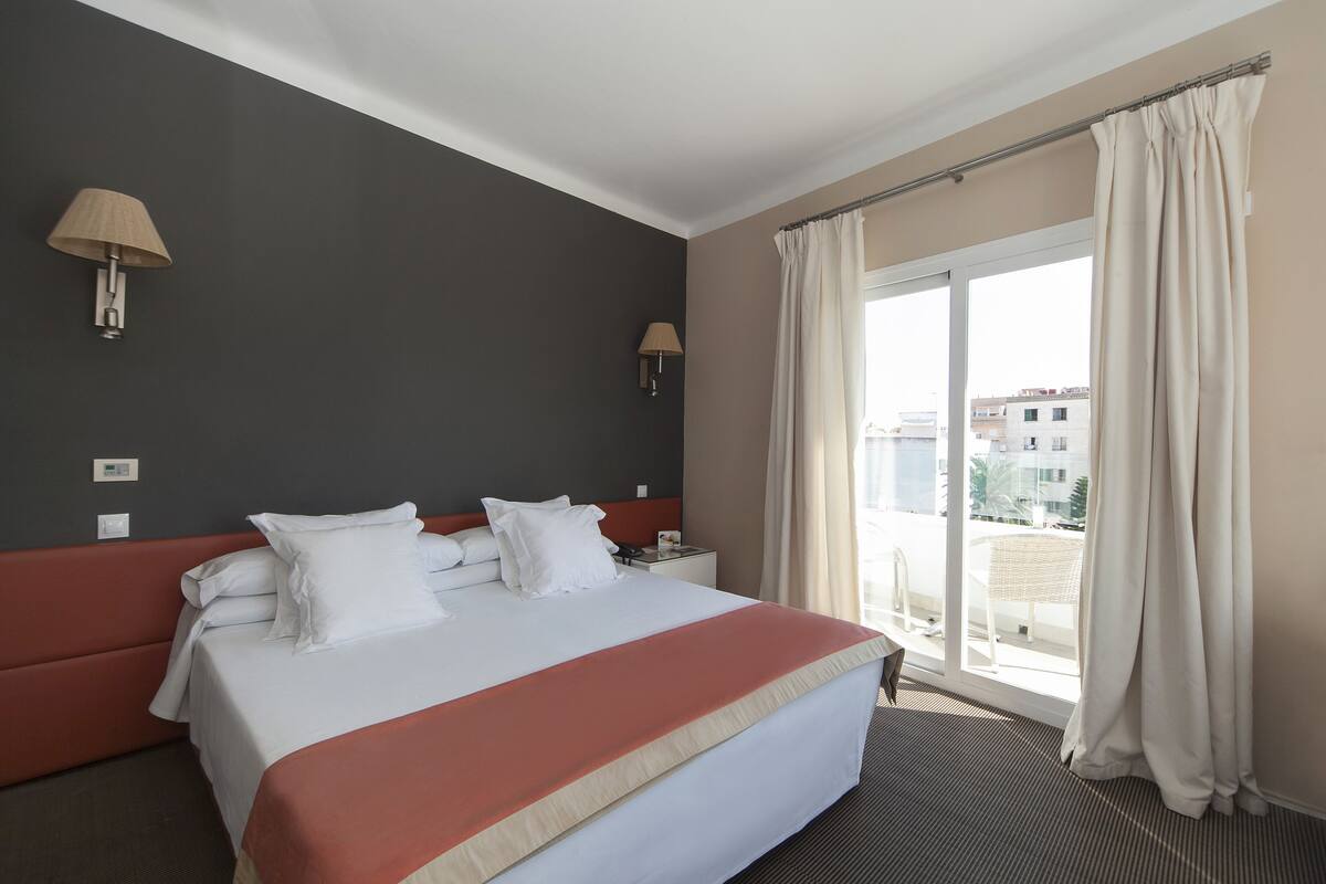 Ein Hotelzimmer mit einem Bett, zwei Wandlampen, einem Fenster mit Vorhängen und Blick auf Gebäude.