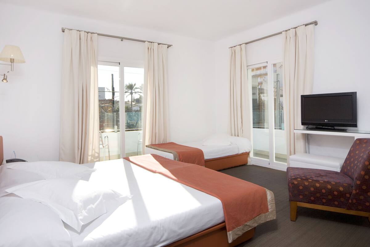 Ein Hotelzimmer mit zwei Betten, einem Fernseher, einem Sessel und großen Fenstern mit Vorhängen.