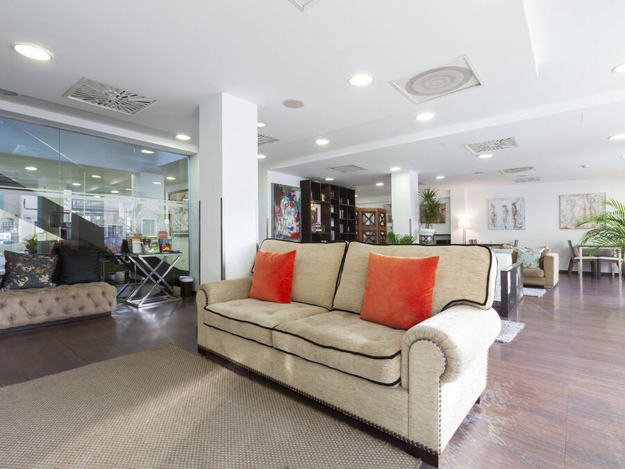 Un vaste espace de vie comprenant un canapé beige, une table basse et une table à manger avec des chaises. On aperçoit des étagères et une plante en pot à l’arrière-plan.