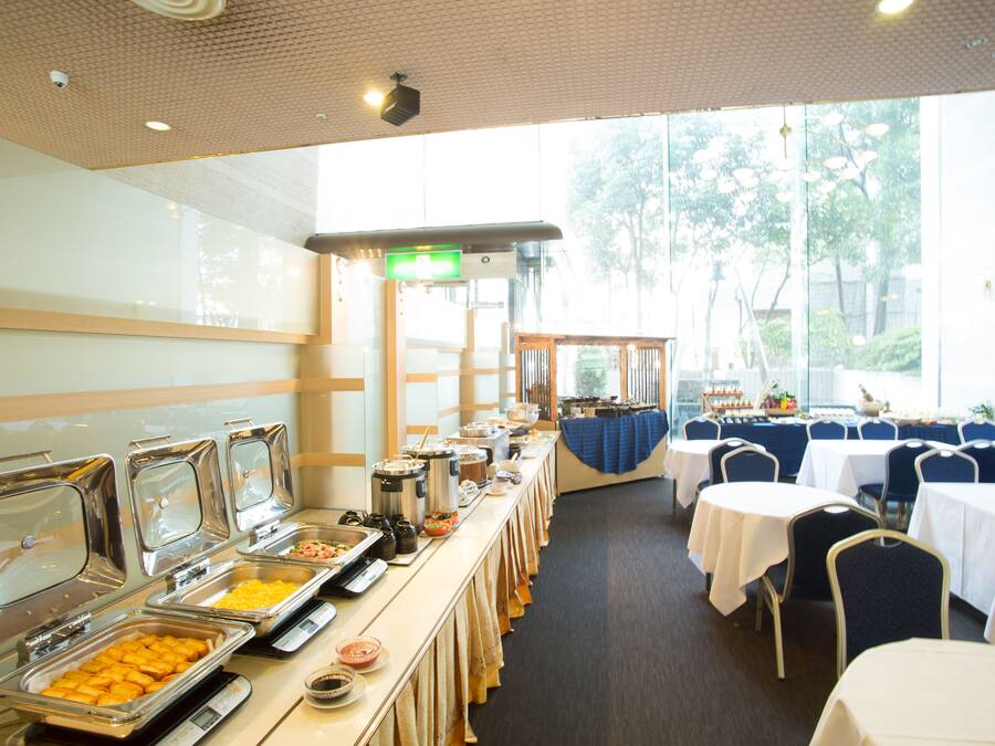 Disposición de un bufé con varias estaciones de comida, mesas y sillas en una sala amplia con grandes ventanales.