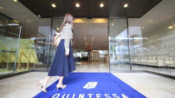 A woman walking on a blue carpet with the word "QUINTESSA" in a modern lobby with glass walls and a seating area.