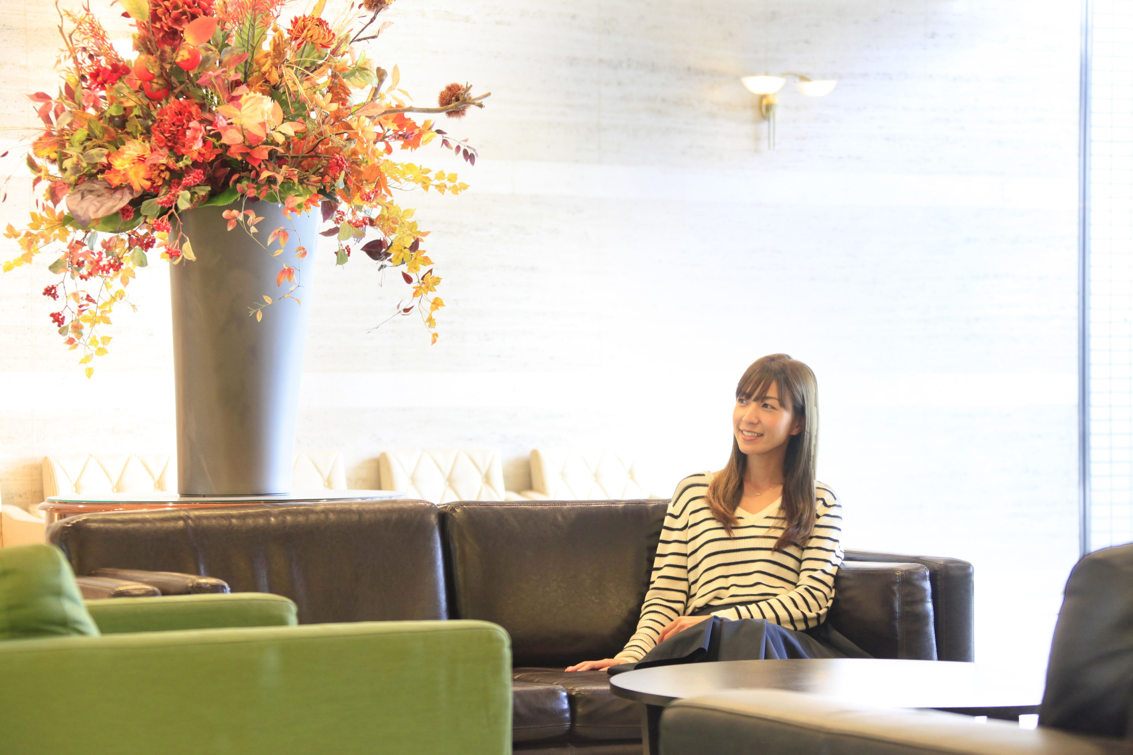 A woman seated on a brown leather couch in a room with a large floral arrangement in a grey vase.