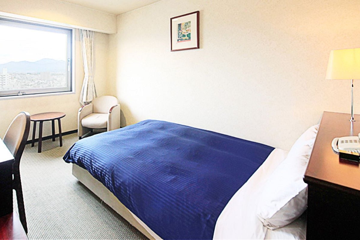 Une chambre d’hôtel comprenant un lit, un bureau, une chaise, une table de chevet avec une lampe, une fenêtre donnant sur un paysage agréable et un tableau encadré au mur.
