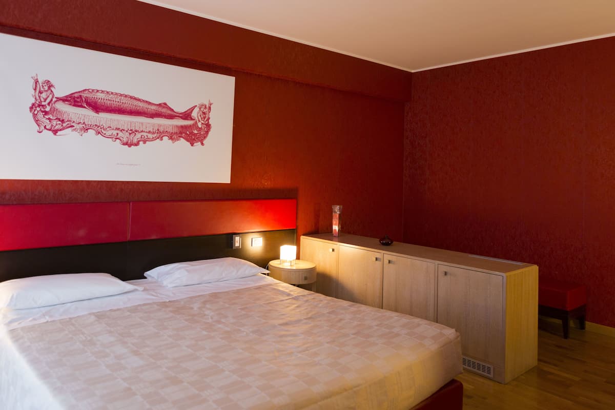 A hotel room with a large bed, a wooden headboard, a bedside table with a lamp, a wooden dresser, and a red accent wall.