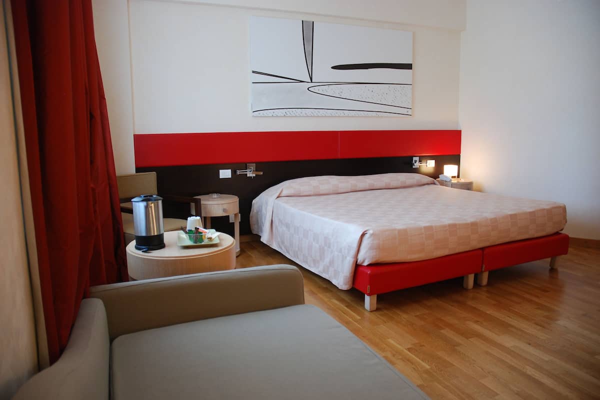 A hotel room with a bed, a bedside table with a lamp, a chair, and a red curtain.