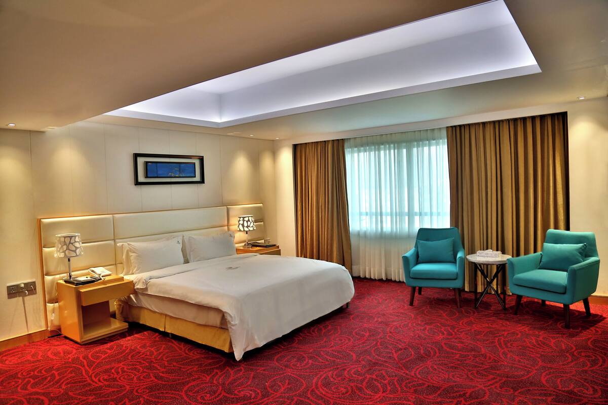 Habitación de hotel con una cama grande, un escritorio con lámpara, dos sillones, una mesita y una ventana con cortinas.