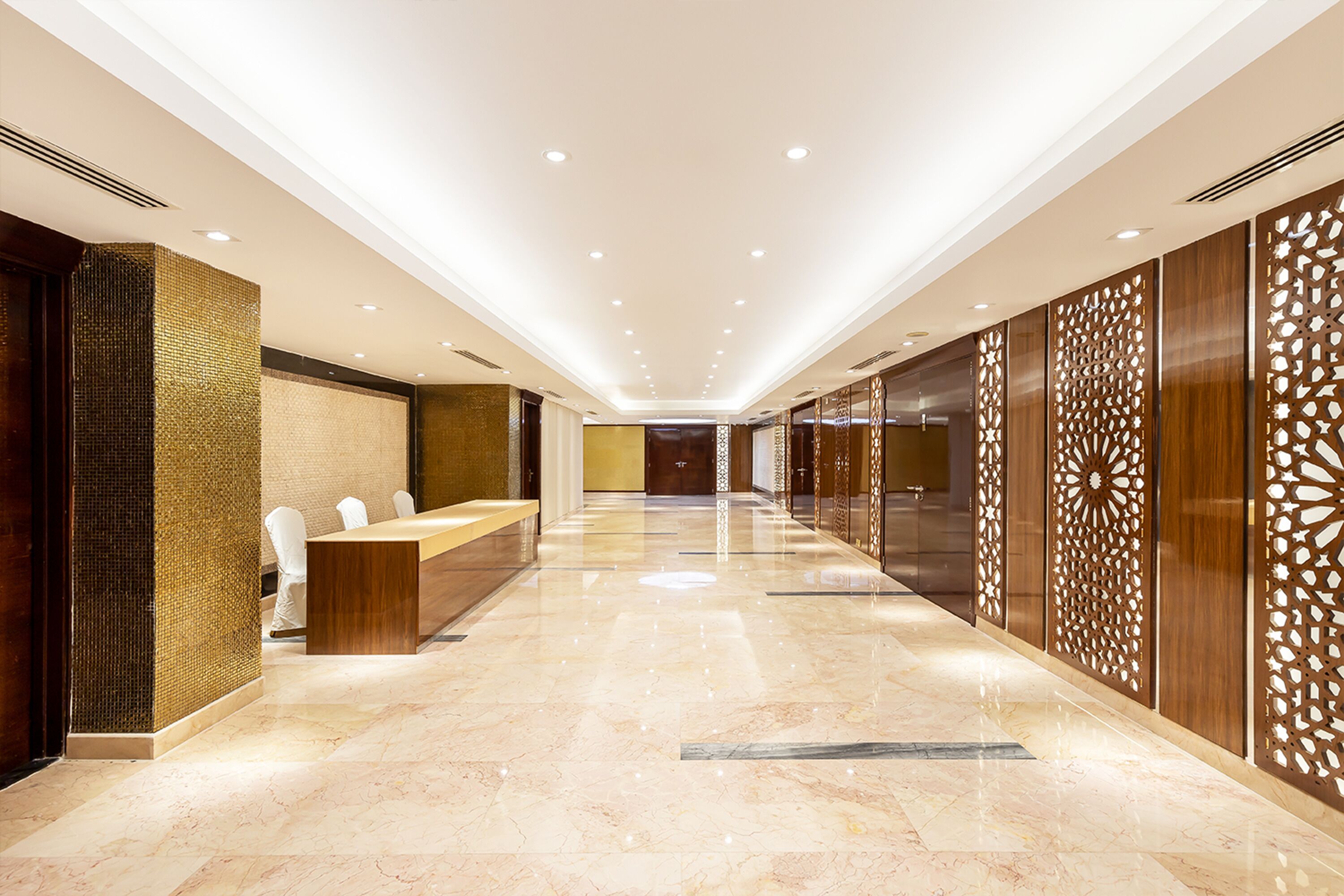 A hotel lobby with a long reception desk, a high ceiling with recessed lighting, and a patterned wall on the right.