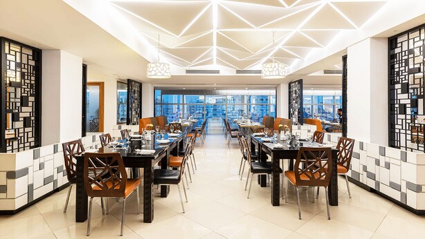 A modern dining area with geometric ceiling lighting, a central aisle, and a view of the city through large windows. - Dhaka Regency Hotel & Resort (Dhaka)