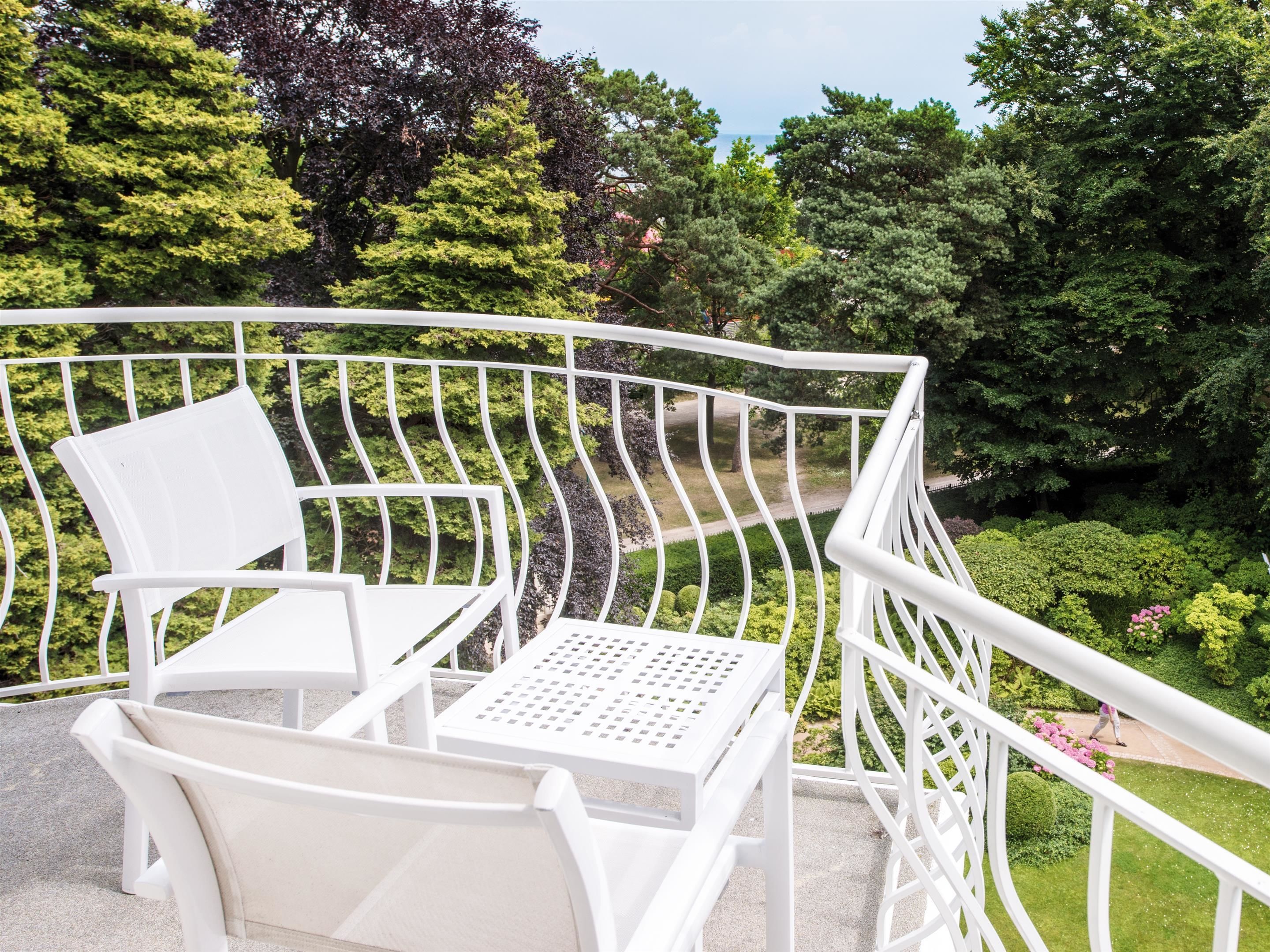 Ein weißer Sitzbereich im Freien mit Tisch und Stühlen, umgeben von einem weißen Metallgeländer, mit Blick auf einen gepflegten Garten mit verschiedenen Bäumen und Sträuchern.