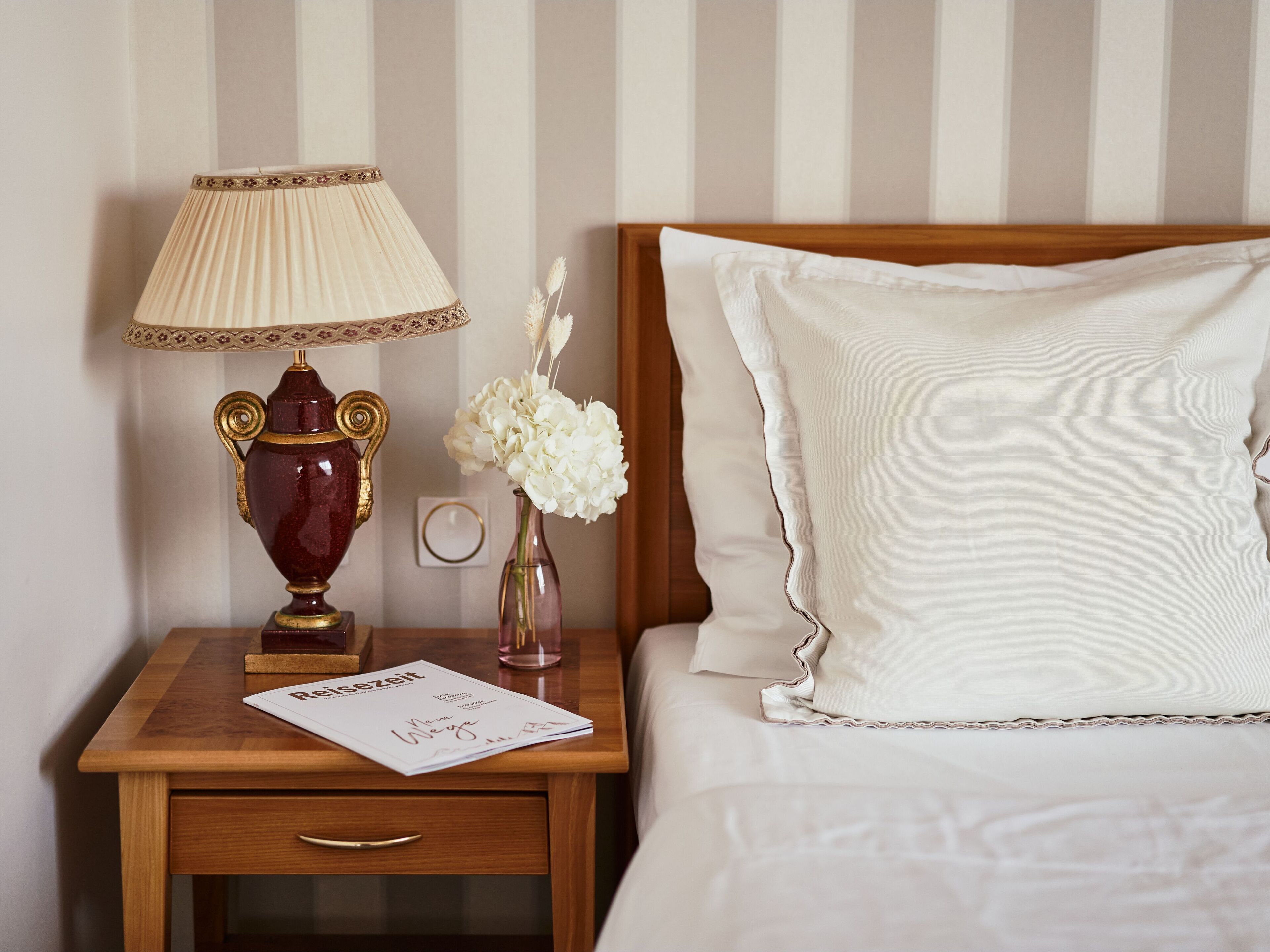 Una cama bien hecha con ropa blanca, una mesita de noche de madera con una lámpara, un florero con flores blancas y una revista sobre la mesita.