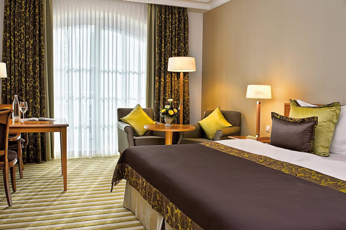A hotel room with a large bed, a desk with a chair, a small table with a lamp, and curtains.