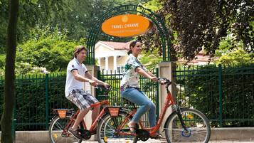 Two people riding bicycles on a path with a green fence and trees in the background.