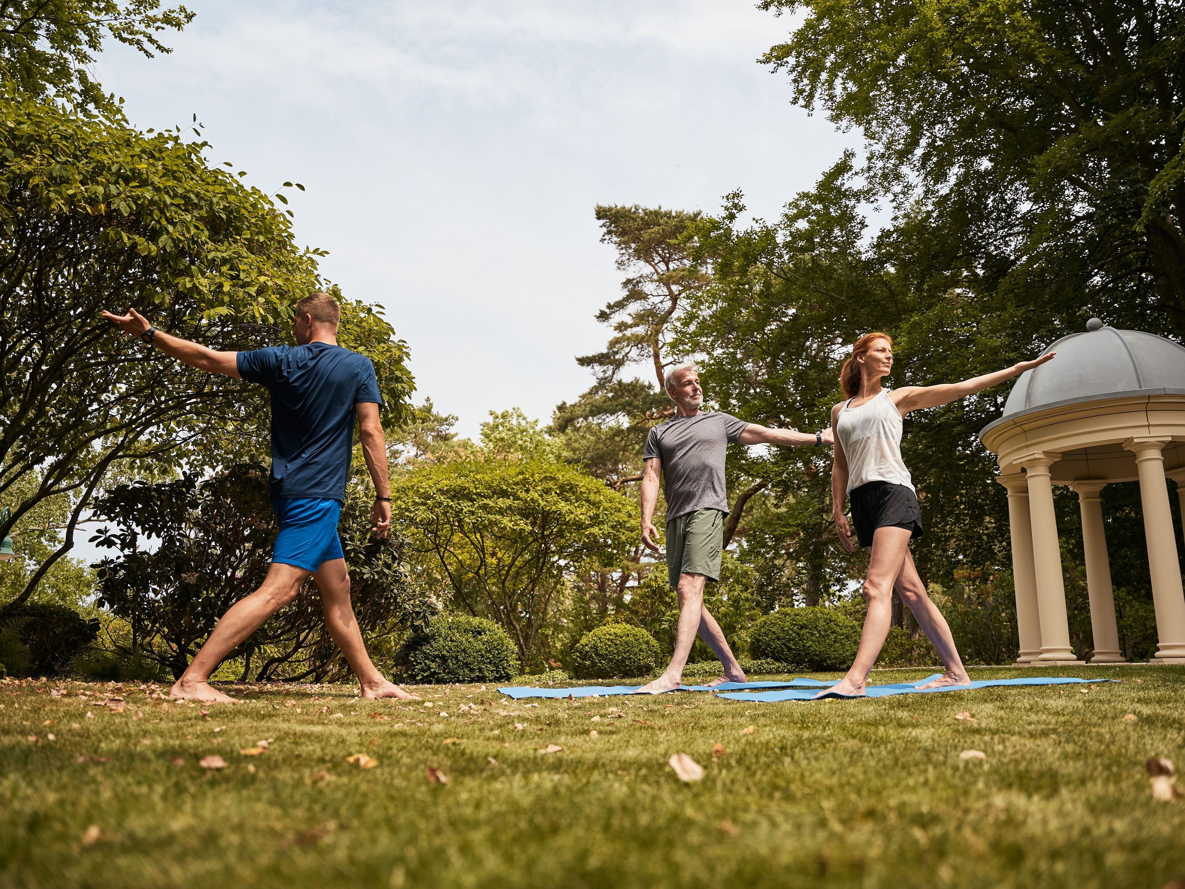 Tres personas practicando yoga en un campo cubierto de césped, con árboles y un pabellón al fondo.