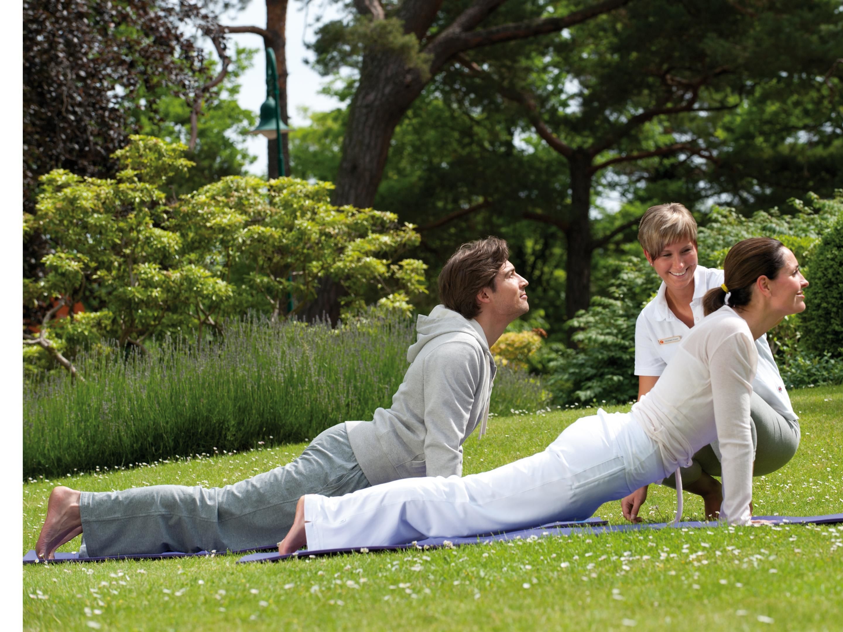 Un grupo de personas realizando un estiramiento de yoga sobre una colchoneta en un parque con árboles y césped.