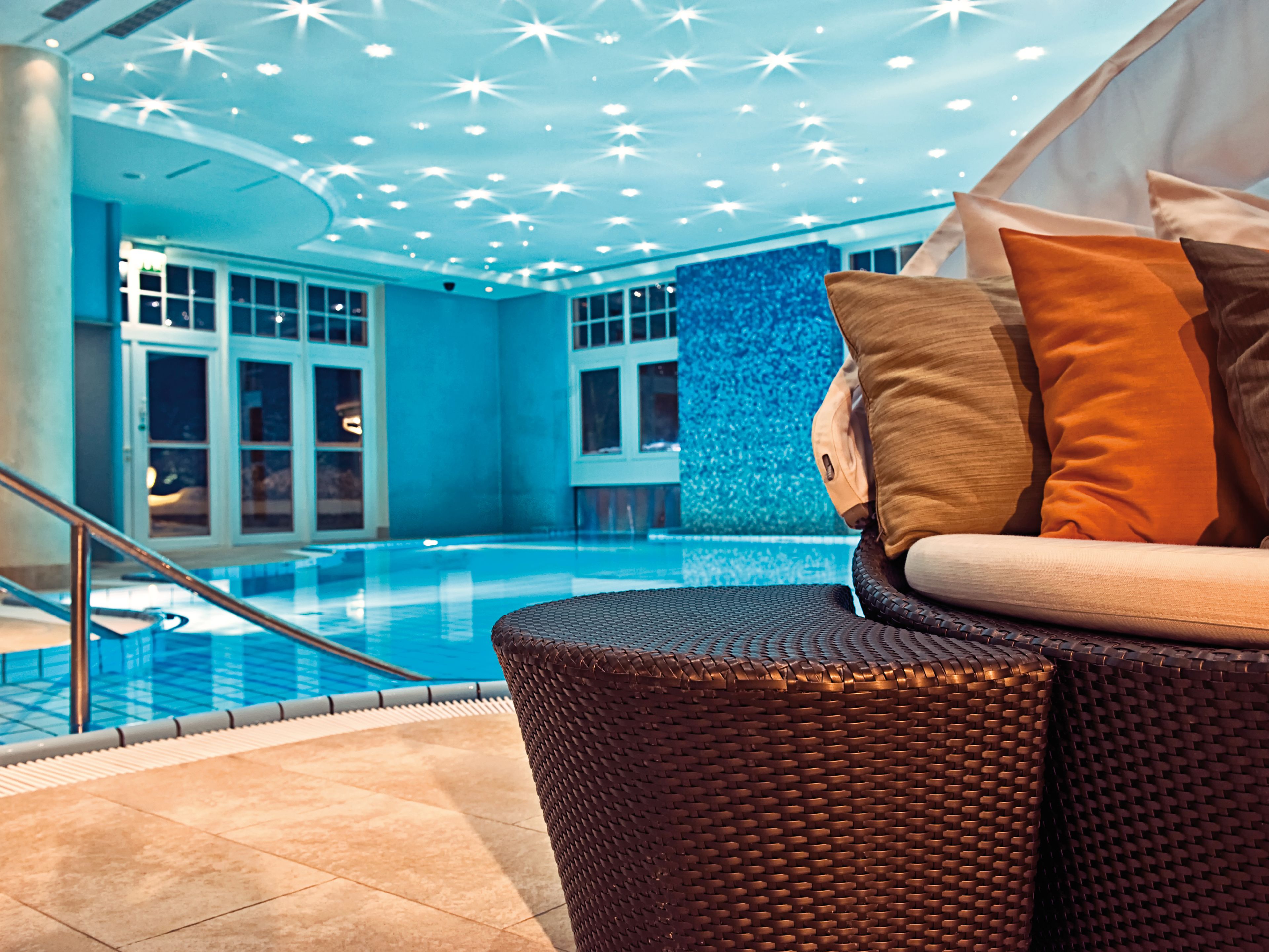 Un espace piscine intérieur moderne, doté d’une paroi en verre transparent, d’un fauteuil de relaxation en osier avec des coussins et d’un mur bleu à motifs texturés.