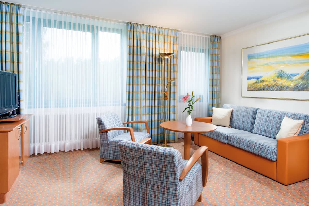 Ein Hotelzimmer mit einer Couch, zwei Sesseln, einem kleinen Tisch mit einer Vase, einem Fernseher und einem Bild an der Wand.