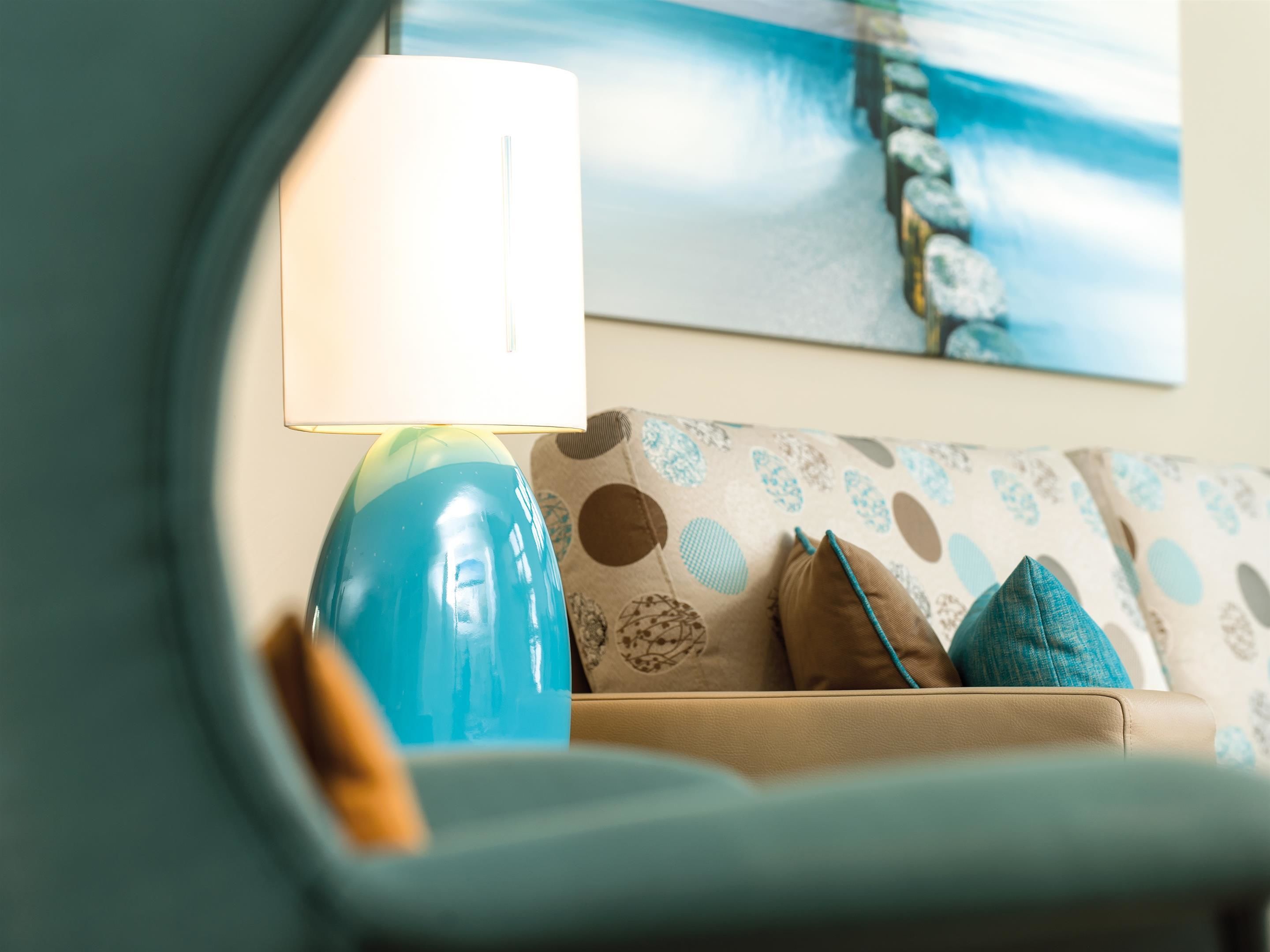 A room with a blue lamp on a beige sofa, a patterned cushion, and a painting of a rocky landscape on the wall.