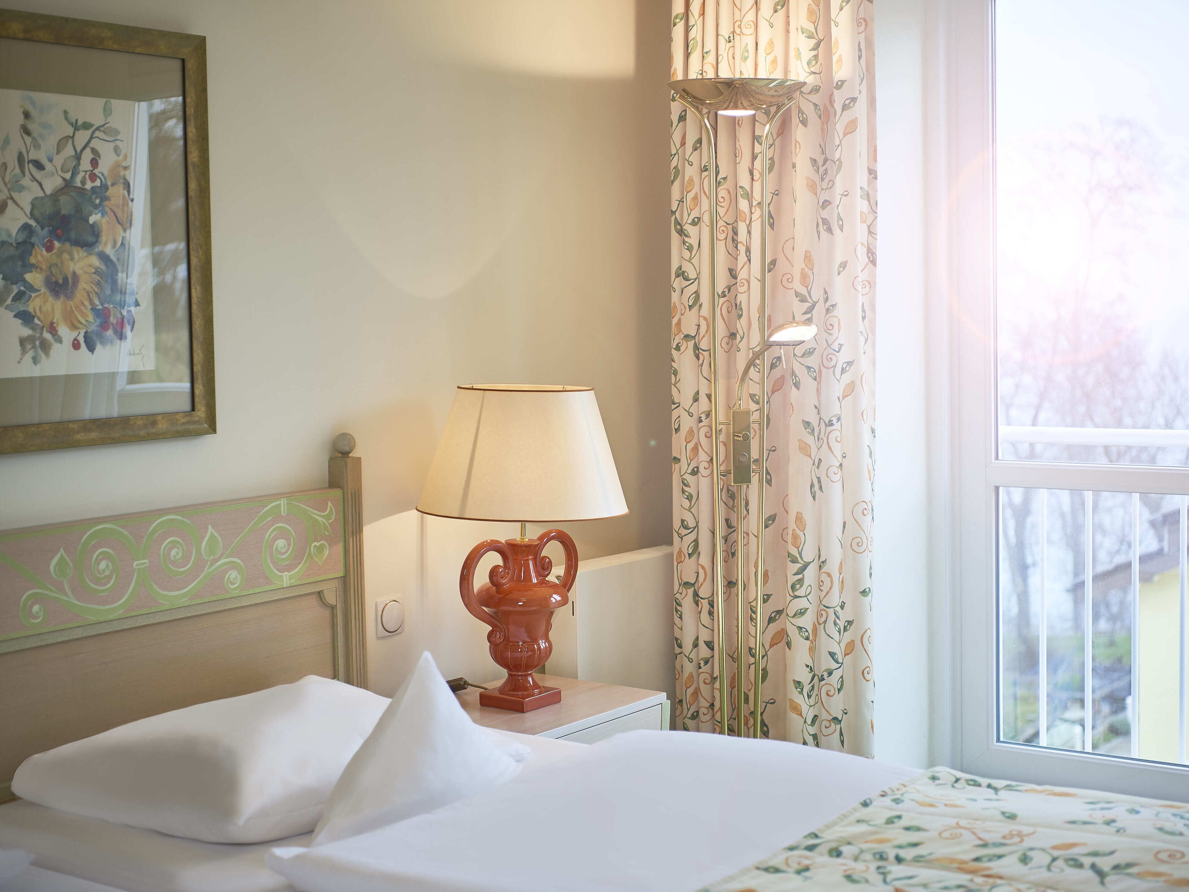 A neatly made bed with white linens, a bedside lamp with a floral design, and a window with curtains.